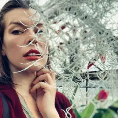 Milla Jovovich, znana z wielu filmów, w zbliżeniu, z roślinami zasłaniającymi część twarzy.