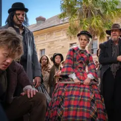 Młody chłopak, jak **the artful dodger**, pochyla się, podczas gdy tłum ludzi w strojach z epoki obserwuje coś poza kadrem.