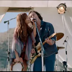 Daisy Jones and the Six: wokalistka i gitarzysta śpiewają razem do mikrofonu, ona trzyma tamburyn.