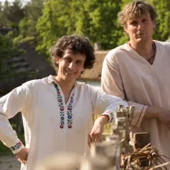 Dwóch mężczyzn w strojach ludowych, jak z filmu "Sami swoi", pozuje na tle wiejskiego krajobrazu.