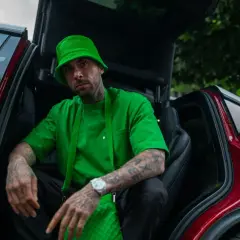 Malik Montana w zielonym kapeluszu i bluzie siedzi w otwartym samochodzie. Jego tatuaże i zegarek przyciągają wzrok, a jego wzrost jest imponujący.