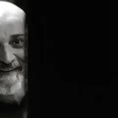 Uśmiechnięty Wojciech Sęp Dudziński, z siwą brodą i łysą głową, wygląda zza ciemnej zasłony.
