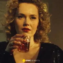 Kobieta z burzą blond loków i czerwoną szminką, pijąca drinka. W tle widać fragmenty scenografii, sugerujące klimat filmu "Jak pokochałam gangstera obsada".