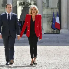 Brigitte Macron młoda, w czerwonej marynarce i czarnych spodniach, idzie za rękę z mężem przed Pałacem Elizejskim.