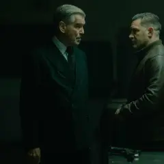 Dwóch mężczyzn w mrocznym pomieszczeniu, jeden w eleganckim garniturze, drugi w skórzanej kurtce. Napięta atmosfera, jakby szykowali się do akcji w mobland season 2.
