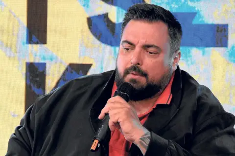 Mężczyzna z brodą i tatuażem na przedramieniu mówi do mikrofonu. Tło to abstrakcyjny wzór. Tylko nie mów nikomu.