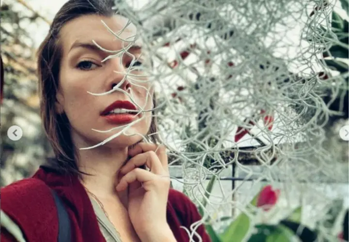 Milla Jovovich, znana z wielu filmów, w zbliżeniu, z roślinami zasłaniającymi część twarzy.