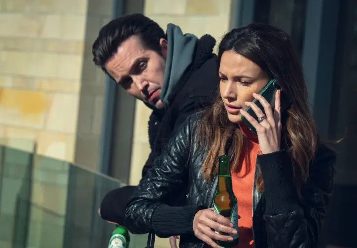 Kobieta rozmawia przez telefon, trzymając piwo. Mężczyzna za nią patrzy z podejrzliwością. Już mnie nie oszukasz.