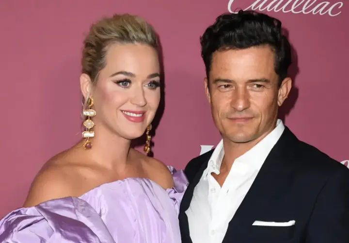 Katy Perry i Orlando Bloom na tle różowej ściany. Ona w fioletowej sukni, on w garniturze.