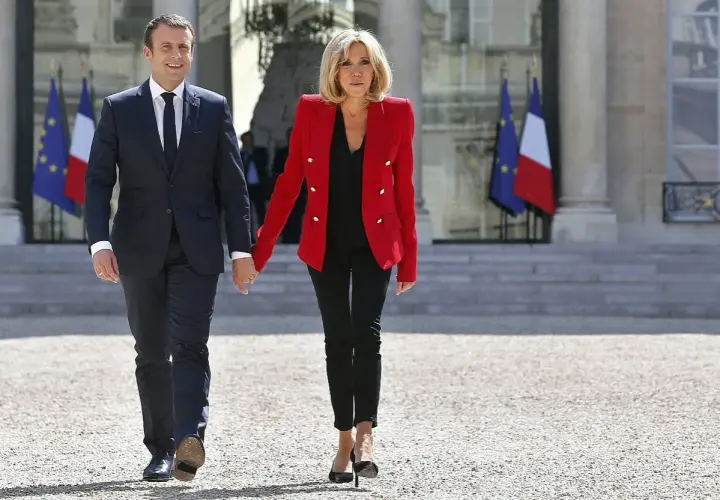 Brigitte Macron młoda, w czerwonej marynarce i czarnych spodniach, idzie za rękę z mężem przed Pałacem Elizejskim.