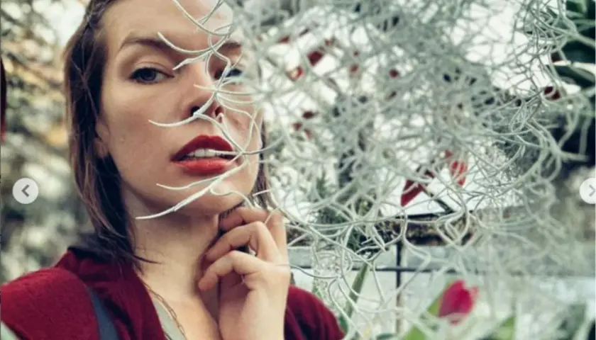 Milla Jovovich, znana z wielu filmów, w zbliżeniu, z roślinami zasłaniającymi część twarzy.