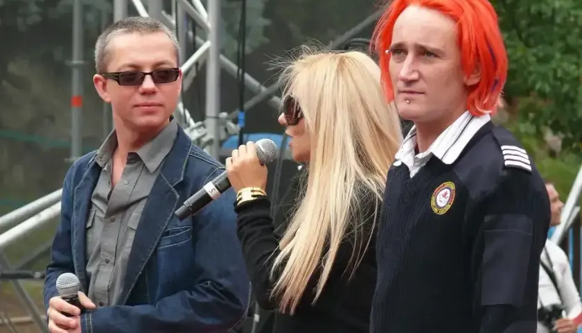 Ich troje wokalistki na scenie, z mikrofonami w dłoniach. Jeden z nich ma ogniste, pomarańczowe włosy.