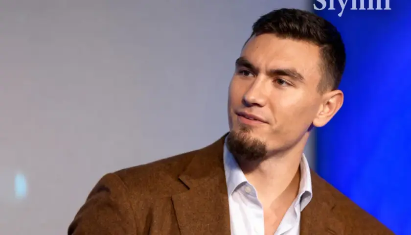 Młody mężczyzna w brązowej marynarce, z zarostem. Conan Kaźmierski wiek to temat dyskusji, ale jego pewność siebie mówi wiele.