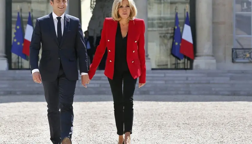 Brigitte Macron młoda, w czerwonej marynarce i czarnych spodniach, idzie za rękę z mężem przed Pałacem Elizejskim.