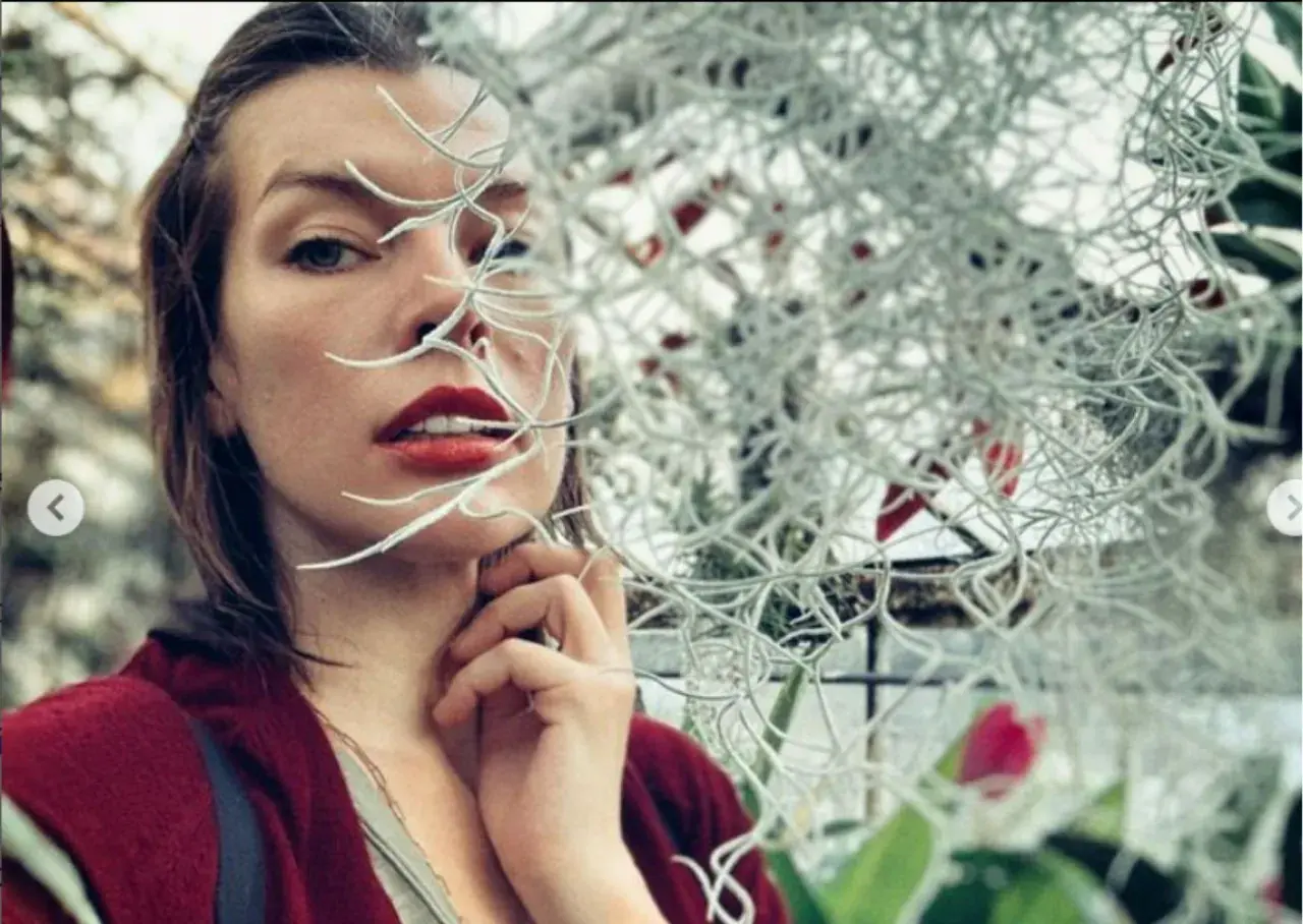 Milla Jovovich, znana z wielu filmów, w zbliżeniu, z roślinami zasłaniającymi część twarzy.