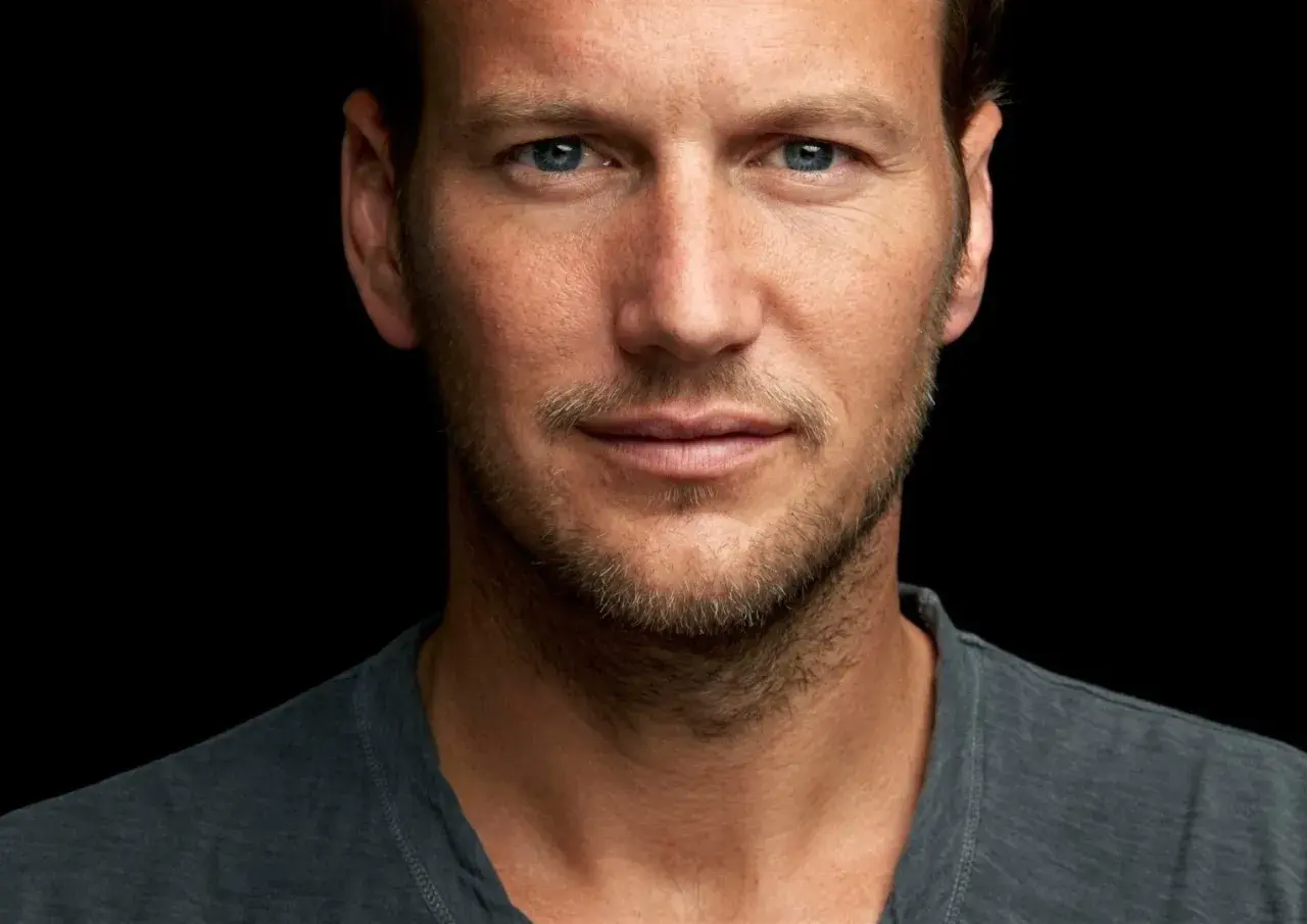 Patrick Wilson, znany z wielu filmów, z niebieskimi oczami i lekkim zarostem, patrzy wprost na widza.