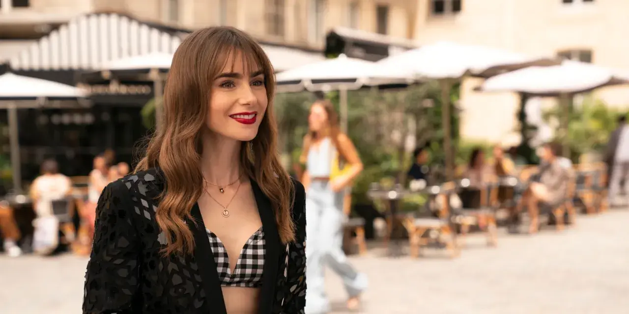 Emily w Paryżu, uśmiechnięta, w stylowej stylizacji, na tle paryskiej kawiarni.
