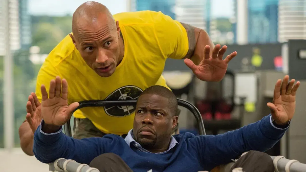 Dwayne Johnson w żółtej koszulce i Kevin Hart w niebieskim swetrze, w scenie akcji z jednego z ich filmów.