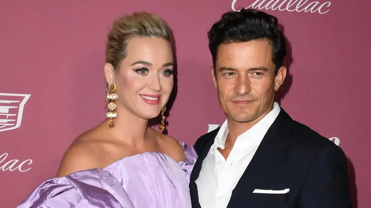 Katy Perry i Orlando Bloom na tle różowej ściany. Ona w fioletowej sukni, on w garniturze.