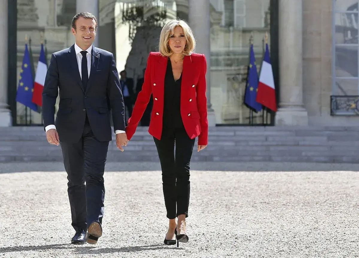 Brigitte Macron młoda, w czerwonej marynarce i czarnych spodniach, idzie za rękę z mężem przed Pałacem Elizejskim.