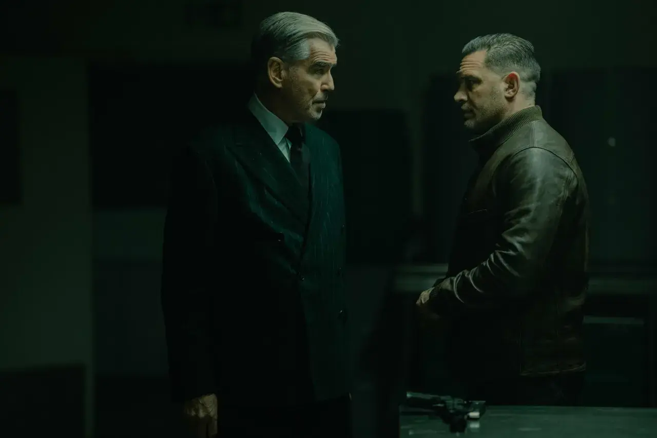 Dwóch mężczyzn w mrocznym pomieszczeniu, jeden w eleganckim garniturze, drugi w skórzanej kurtce. Napięta atmosfera, jakby szykowali się do akcji w mobland season 2.