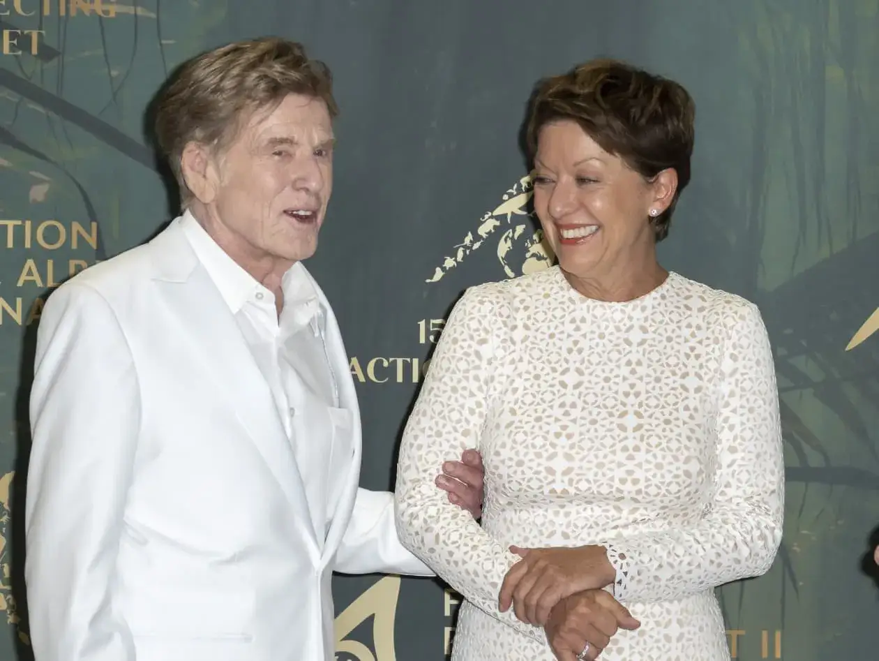 Robert Redford z żoną, oboje uśmiechnięci, pozują do zdjęcia. On w białym garniturze, ona w koronkowej sukience.