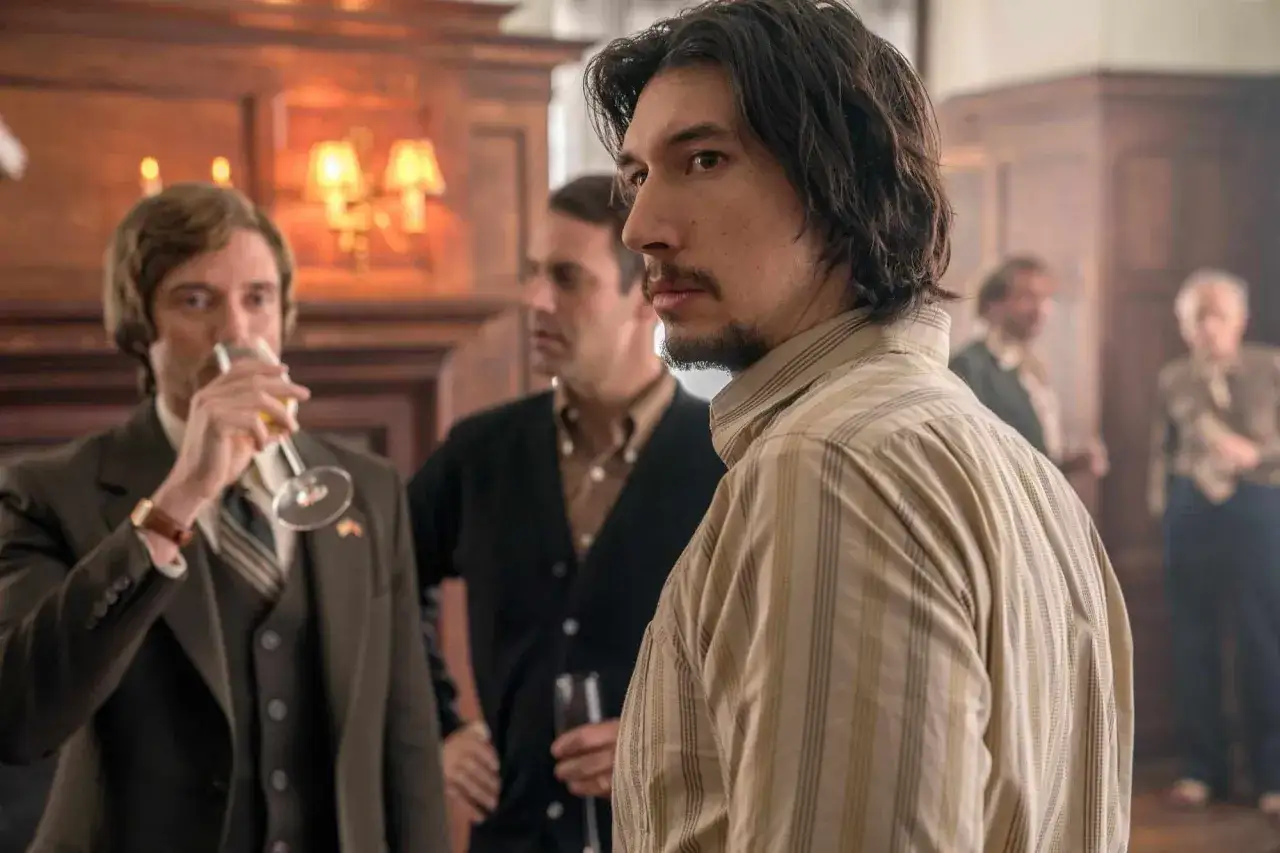 Adam Driver w filmach, w eleganckim stroju, rozmawia z mężczyznami na przyjęciu.