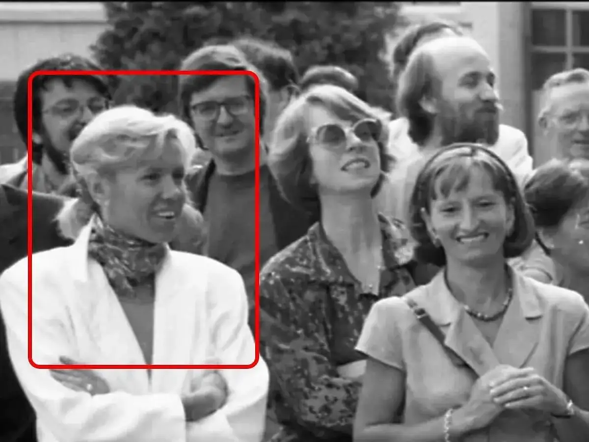 Brigitte Macron młoda, w białej marynarce i z apaszką na szyi, uśmiecha się do obiektywu. W tle widać innych ludzi.
