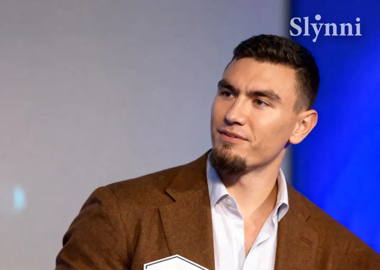 Młody mężczyzna, Conan Kaźmierski, w brązowej marynarce i białej koszuli, patrzy w bok.