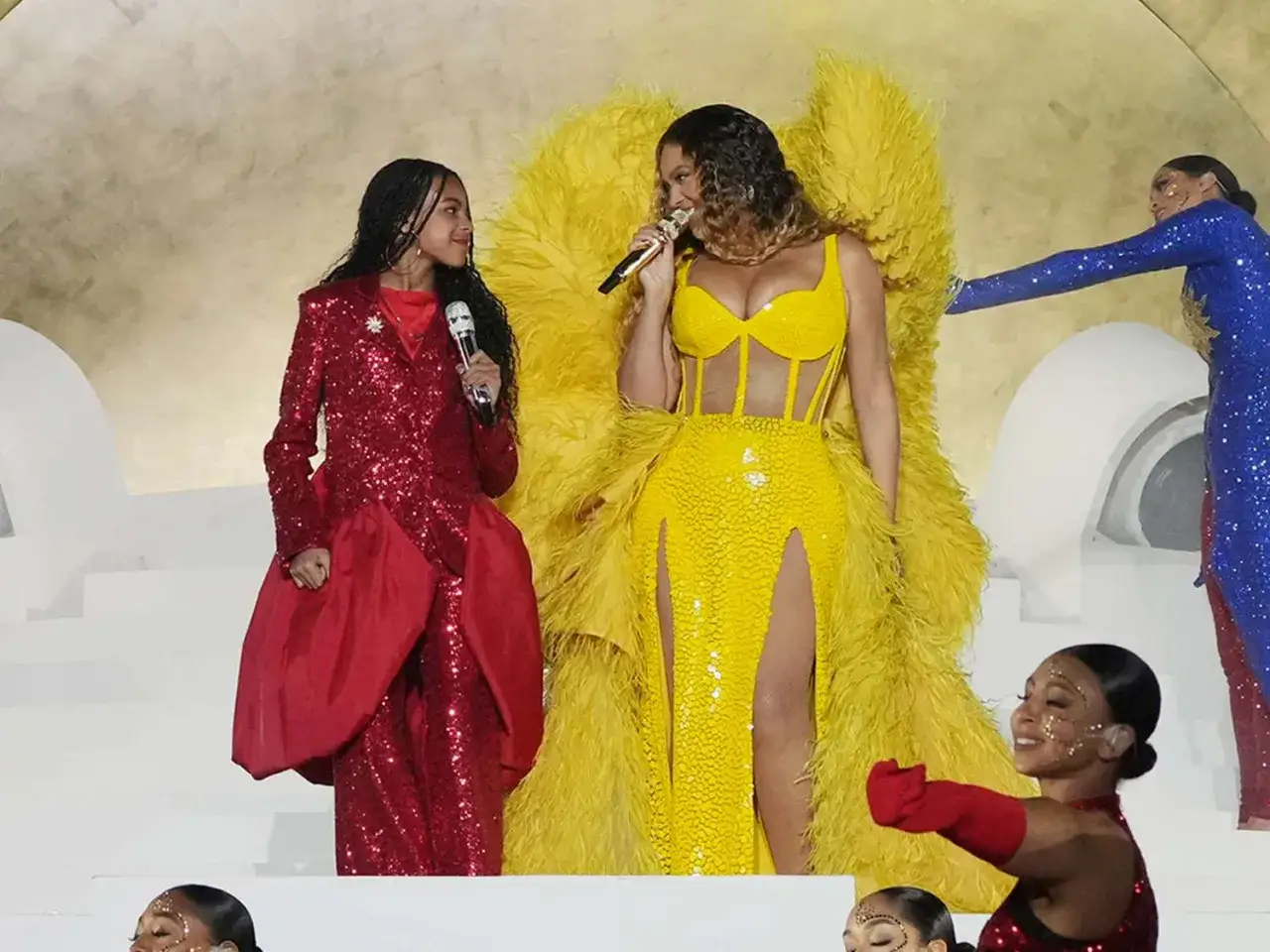 Blue Ivy Carter w czerwonej, błyszczącej kreacji obok Beyoncé w żółtym stroju na scenie.