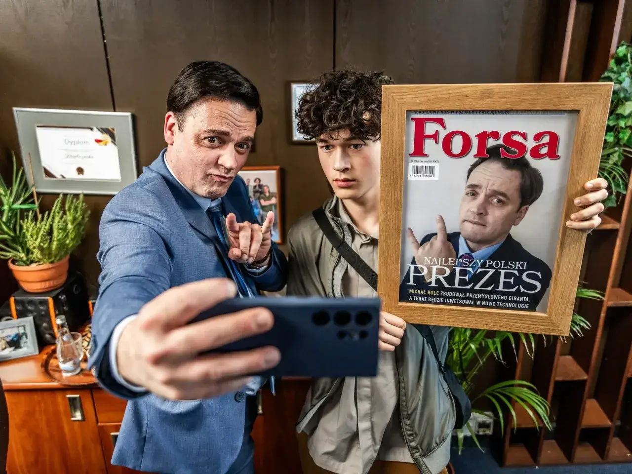 Dwóch mężczyzn w biurze, jeden robi selfie z ramką z okładką magazynu 