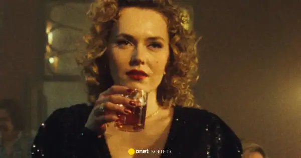 Kobieta z burzą blond loków i czerwoną szminką, pijąca drinka. W tle widać fragmenty scenografii, sugerujące klimat filmu "Jak pokochałam gangstera obsada".